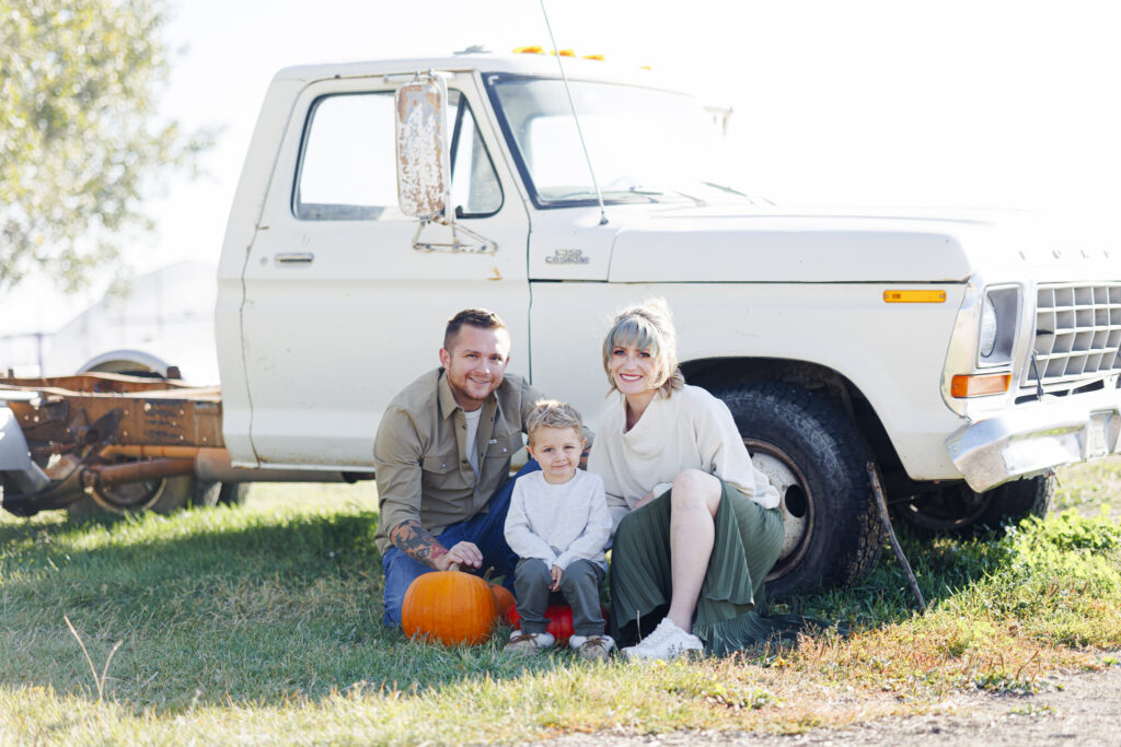 Family of three smiling at camera in front of a white truck with pumpkins around