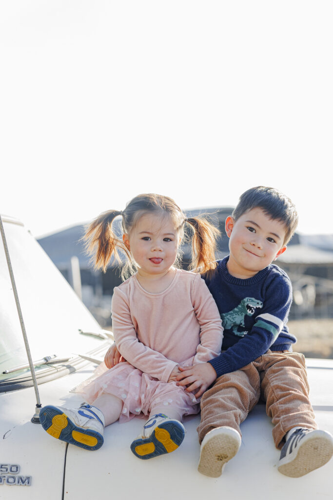 brother and sister sit together on the hood of a white Ford truck