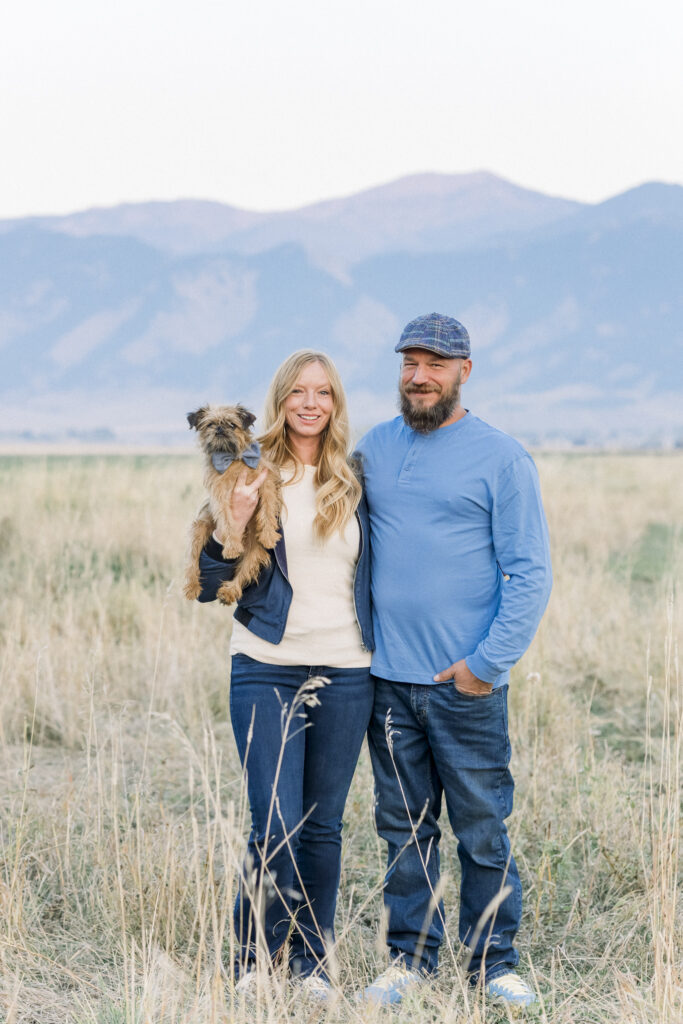 man and woman hold dog wearing blue bowtie in front of Bridger mountains